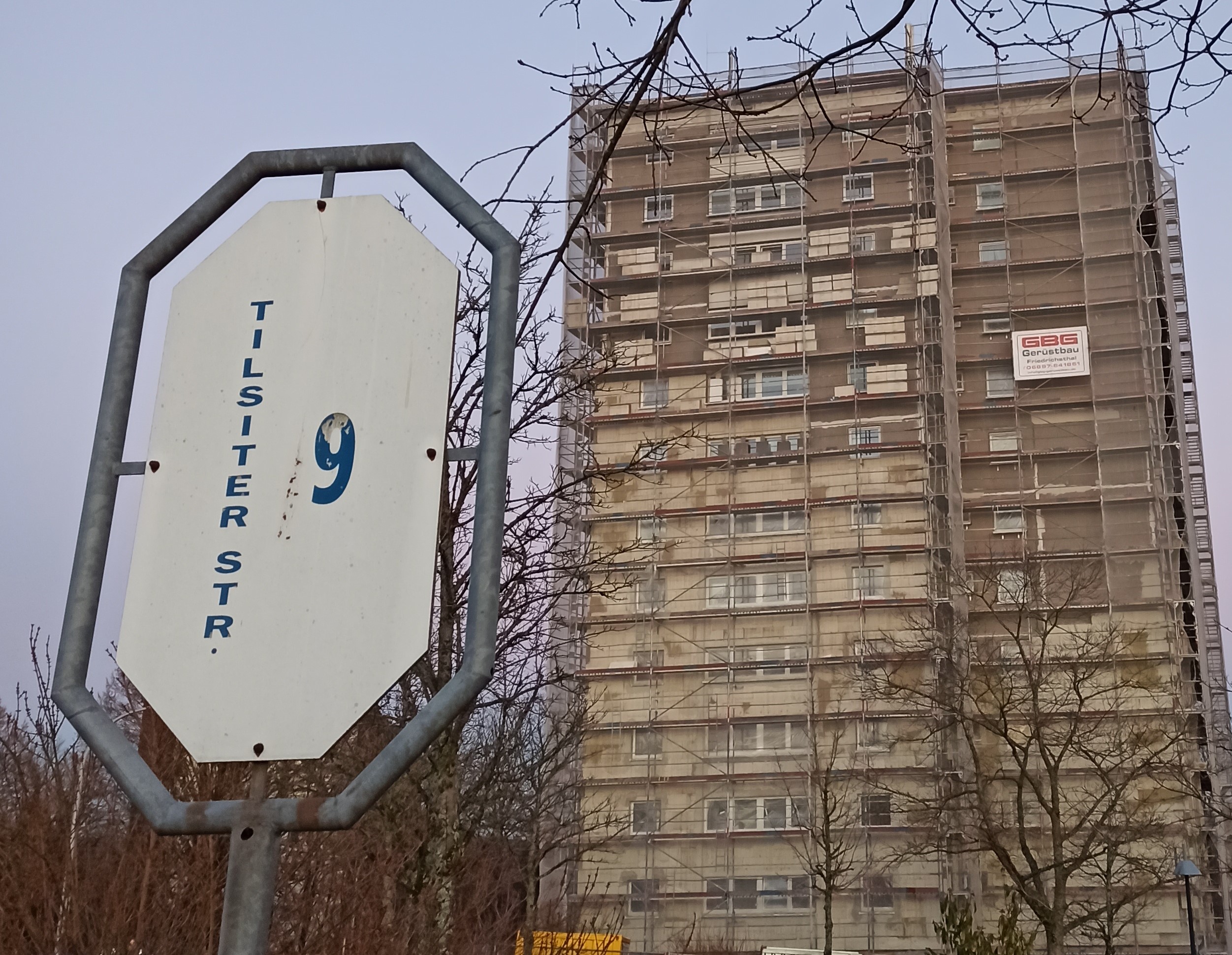 In dem Bild sieht man ein Studentenwohnheim. Im Vordergrund des linken Bildrandes steht ein weißes Schild mit grauem Rahmen, auf dem die Adresse Tilsiter Straße 9 steht. Dahinter sieht man das zwölfstöckige Gebäude, umgeben von einem Gerüst auf Grund von Sanierungsarbeiten. In diesem Haus befinden sich die Studentenwohnheime.